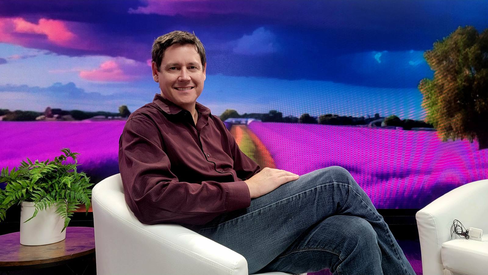 Chris Brickler seated in a studio against a wall-sized image of a lavender field at dusk
